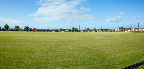 A wide, open community sports field in a suburban neighborhood in Tarneit, Australia, surrounded by houses. Flat green grass for cricket, football, and other outdoor activities. Local recreation area.