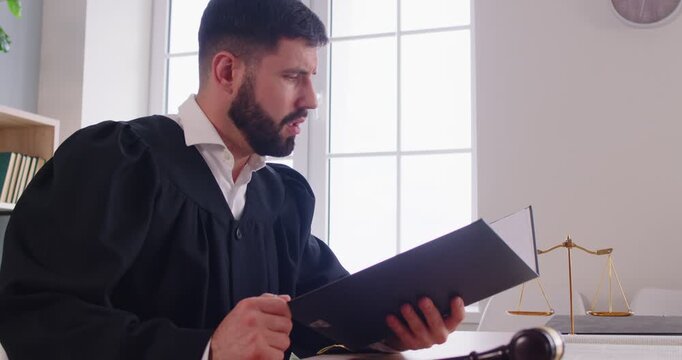 Confident young man judge wearing judicial robe sitting at the desk on workplace reading out the verdict in courtroom. Male justice working indoors. Legal law concept and justice concept. 4k video.