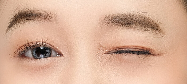 Close-up of a woman winking, showing one open eye with blue-gray contact lens and long lashes, highlighting smooth skin and natural makeup in soft lighting. - Powered by Adobe