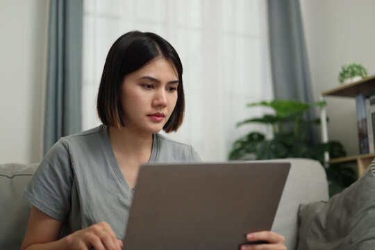 Front view, Young Asian woman in casual clothes using laptop while sitting on sofa in cozy living room. Work from home, Remote working.