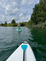Fototapeta premium Serene POV point-of-view shot from a white SUP board gliding across calm lake or river waters. An active man standing on their knees is paddling in front. Active lifestyle getaway for group or couple 