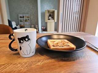 Banana cheese toast and green tea in an adorable cat mug are served on a wooden table. A cozy and homey image of a simple, delicious breakfast in a modern Japanese home.
