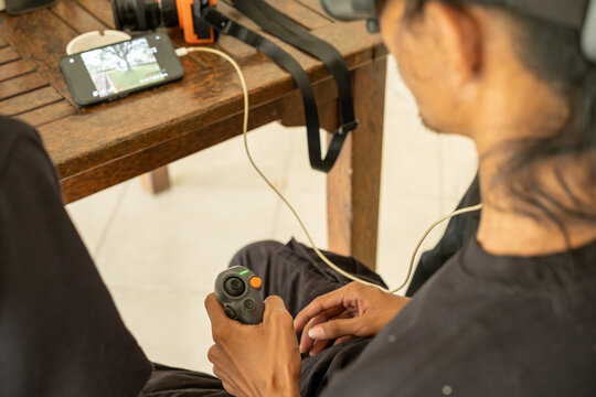Man operating a drone using handheld remote controller while monitoring live video feed on smartphone, indoor setup with wooden table, technology and control concept.