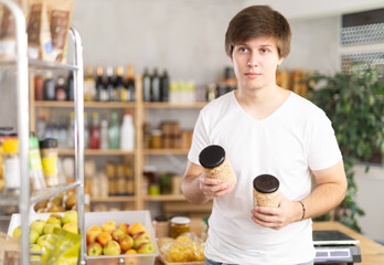 Fototapeta premium Young man store visitor chooses can of beans, hold and view glass jar package. Grocery shop, trading floor, shelf with product items in background.