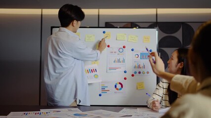 Young businessman conference speaker passionately explains business project strategy using a whiteboard, engaging a diverse team in collaborative brainstorming within a contemporary office environment