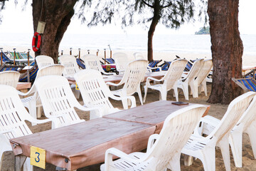 Empty white chairs plastic and tables set up for seaside on beach dining experience under shade of trees with view of ocean and mountains with bright sunlight is background.