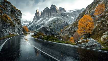 A winding, wet road leads through a dramatic mountain pass, surrounded by jagged peaks with patches of snow and vibrant yellow autumn trees.