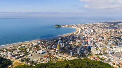 Aerial view of Guaratuba city and beach coastline with calm ocean and buildings under a partly cloudy sky.