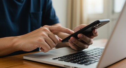 Close up of a person s hands using a smartphone while working on a laptop computer at a wooden desk