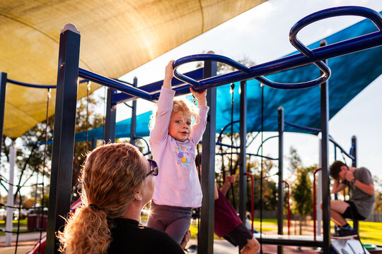 Mum helping toddler girl with curly hair play at park playground on autumn day