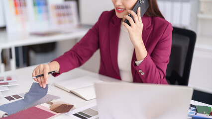 Asian adult woman interior architect selects material samples discusses client project via mobile phone modern office desk
