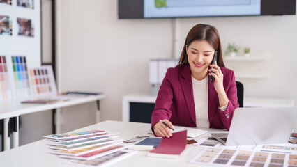 Asian adult female interior designer discuss home decor with client on phone selecting color material samples in modern design studio office