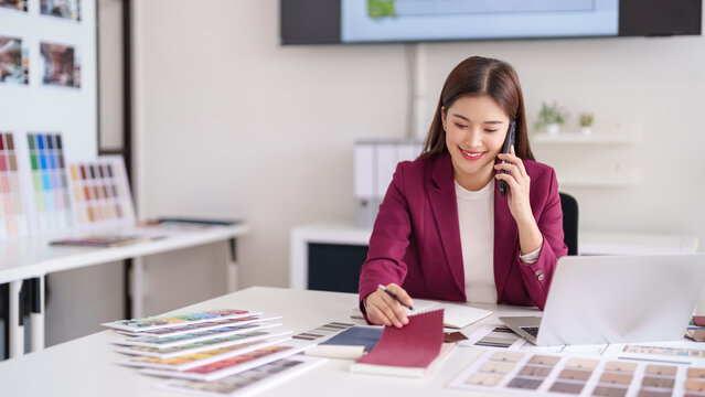 Asian adult female interior designer discuss home decor with client on phone selecting color material samples in modern design studio office