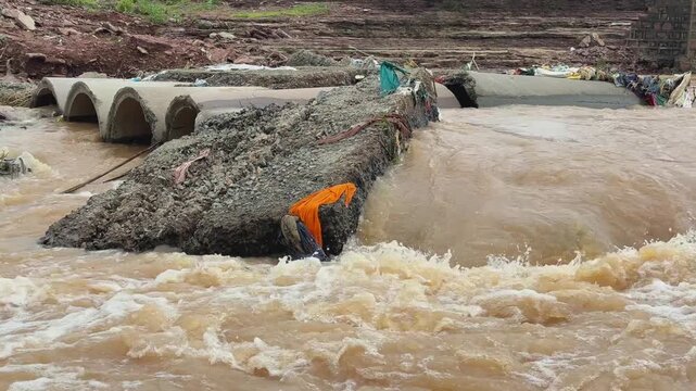 Static shot showing a heavly damaged culvert or a small bridge partially submerged by the powerful, muddy brown water during the flooding