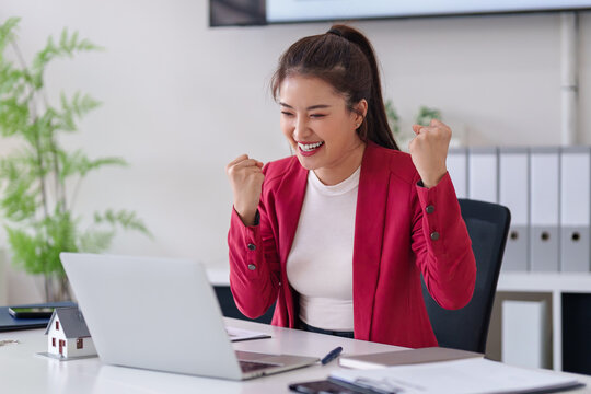 Asian adult woman real estate agent celebrates major sales success closing deal using laptop computer at modern office desk property investment achievement