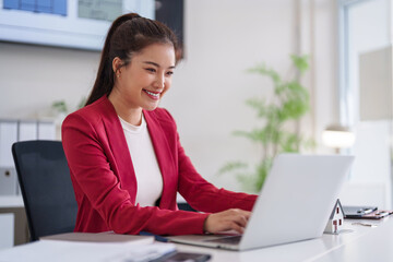 Asian adult business professional smiles while working laptop at modern office desk selling real estate home insurance property investments financial planning