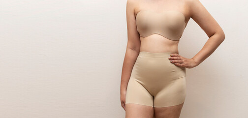 A woman in nude shapewear stands confidently with hands on hips, showcasing body positivity and self-acceptance in neutral space.  © KingKongThePitbull