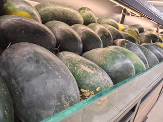 Watermelons in Supermarket Display. A wide shot of fresh, dark green watermelons arranged in neat rows on a refrigerated shelf, seen through a glass partition.