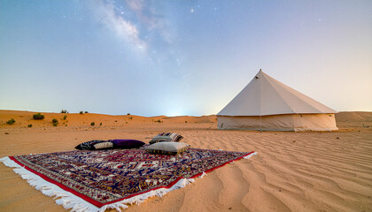 The image shows a cozy campsite in the desert at sunset or sunrise. In the center of the composition, a traditional oriental rug with a bright pattern lies on the sand, with several pillows scattered 