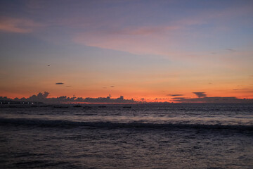 Golden sunset paints the sky over Kuta Beach, Bali, blending warm orange and purple hues above calm ocean waves, creating a serene and breathtaking view along the horizon of this tropical coast.