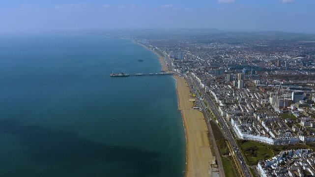 4K drone panorama of Brighton seafront: Palace Pier reaching into the English Channel, i360 and West Pier ruins along a sweeping urban coast, beach and skyline fading toward the South Downs.