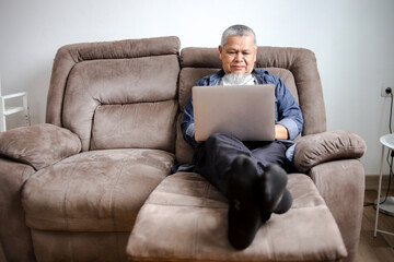 Fototapeta premium Senior Man In Casual Clothing Using Laptop While Sitting On Sofa At Home