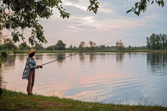 woman in a straw hat fishing on the bank of a serene lake.