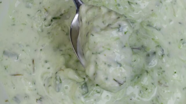 scooping yogurt garlic dip with spoon showing swirl motion in thick white turkish Cacik style sauce with grated cucumber and herbs concept of ethnic cuisine, culinary blog, homemade sauces