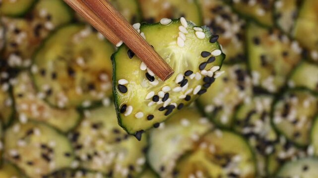 close up of chopsticks lifting soy-marinated cucumber slice with black and white sesame seeds from traditional korean oi muchim dish on textured background
