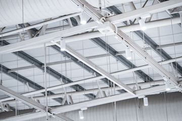 White LED spotlight lamps hanging on the ceiling frame under steel roof truss structure with air duct pipe system nearby. Lighting design installation in modern warehouse or wide span building