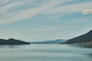 Serene panoramic view of a vast lake with forested mountains under a cloudy blue sky, featuring a distant small boat and a quiet horizon