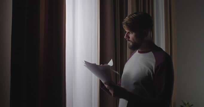 A bearded European man comes to the window holding sheets of paper in his hand. When he sees something frightening or shocking, he drops them