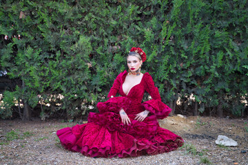 Fototapeta premium Young, beautiful, blonde woman in red flamenco dress, flowers in her hair and gold jewelry, performing flamenco poses sitting on the ground with green plants in the background. Concept dance, art.