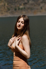 A girl in an orange dress on the shore of a pond, she has long hair falling down her back. Rocks...