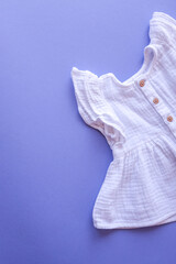 White baby dress with wooden buttons on a light purple background, soft fabric texture, flat lay minimalistic composition, children’s clothing concept