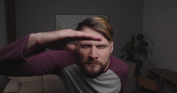 Close-up of a funny, bearded guy looking into the camera, a mirror, or a window, making funny agressive face,places his palm over his eyes, peering into the distance