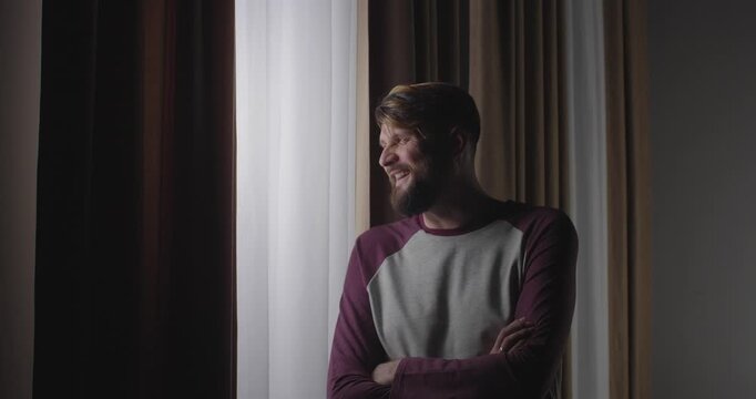 Blonde bearded European  man standing near a window with curtains, looking out and laughing maliciously at what he sees