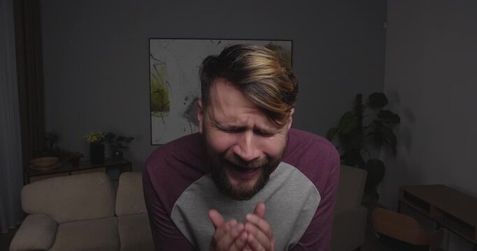 Close-up of a bearded blond man crying funny, stretching out his hands to the camera, standing in a room at home.