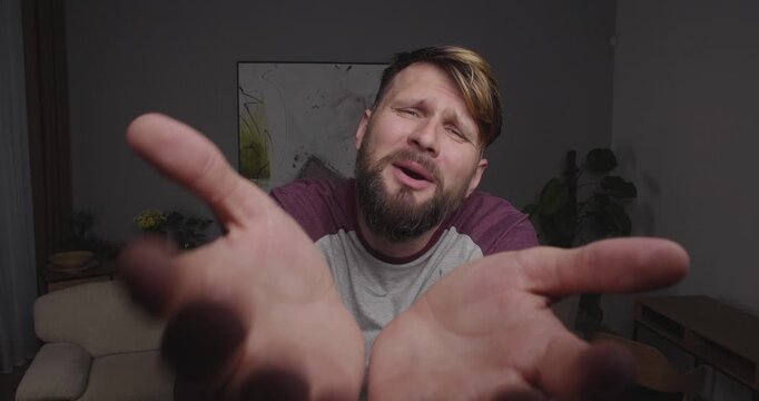Close-up of a bearded blond man crying funny, stretching out his hands to the camera, standing in a room at home.