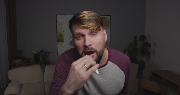 Close-up of a European man, blond and bearded, brushing his teeth in the room looking in the mirror or window. Seeing something  shocking him,  opening his mouth and dropping the brush.