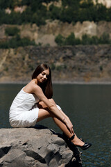 A girl is sitting on the shore of a lake on a large stone, the water in the lake is blue