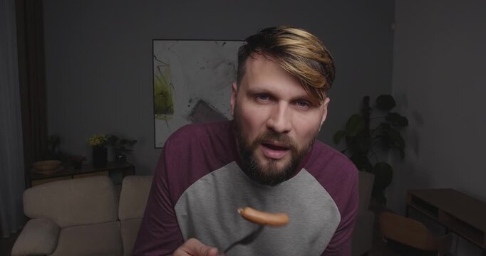 Close-up of a bearded blond man funny eating sausage  looking to the camera, window or tv screen, standing in a room at home with bizarre face, drops his fork scared.