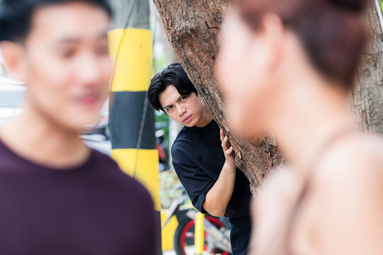Suspicious and obsessed young man hides behind a tree spying on smiling ex-girlfriend laughing closely with new boyfriend during meetup.