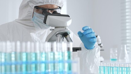 A male doctor in a protective suit and gloves uses a microscope while analyzing samples, conducting...
