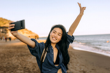 Chinese woman taking a selfie at the beach during sunset while enjoying a carefree moment by the...
