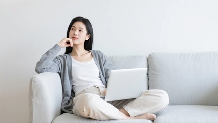 Naklejka premium A thoughtful and focused young woman working on her laptop while sitting comfortably on a gray sofa, portraying a modern and flexible remote work lifestyle.