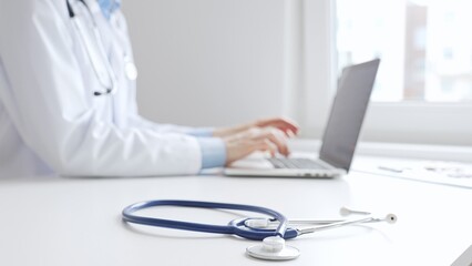 Closeup of blue stethoscope is lying on the table near female doctor working on laptop, highlighting digital tools transforming healthcare diagnostics and patient documentation. Medicine concept