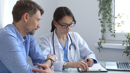 Female doctor consulting with patient in medical office using laptop and filling out paperwork to provide healthcare advice and support, discussing treatment options in a professional setting. Medical