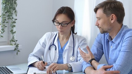 Female doctor consulting with patient in medical office using laptop and filling out paperwork to provide healthcare advice and support, discussing treatment options in a professional setting. Medical