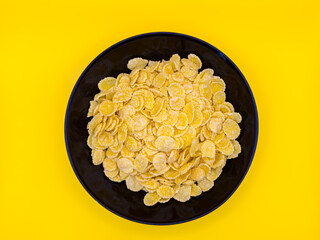 A bowl of yellow corn flakes. The flakes are scattered all over the bowl. The bowl is black in color Corn serial on a yellow background, copy space, top view, flay lay